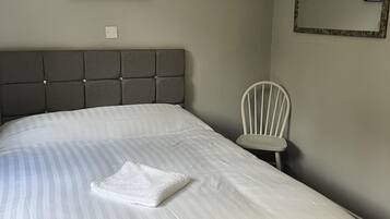 Un lit bien fait, avec une couvre-lit à rayures et une serviette blanche pliée, une tête de lit rembourrée grise, une chaise blanche et des œuvres d’art encadrées accrochées aux murs.