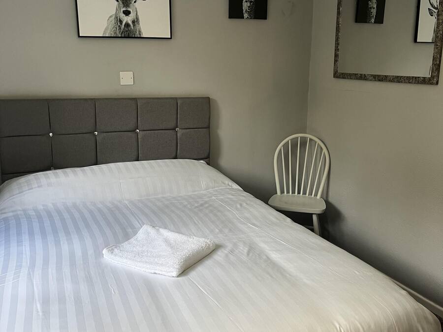 Un lit bien fait, avec une couvre-lit à rayures et une serviette blanche pliée, une tête de lit rembourrée grise, une chaise blanche et des œuvres d’art encadrées accrochées aux murs.