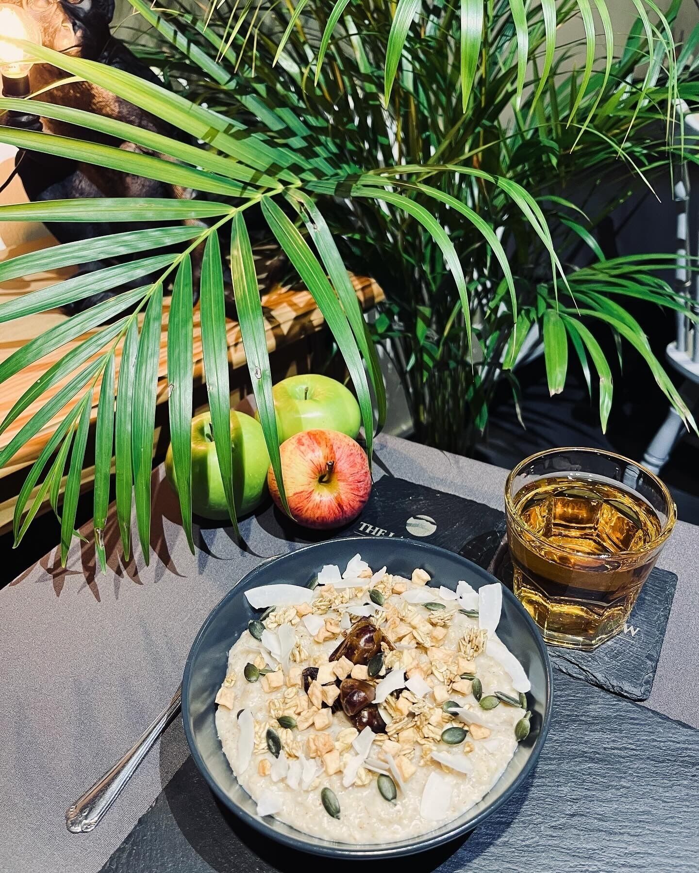 A plate of oatmeal with nuts and seeds, a glass of amber liquid, and apples on a table with a palm leaf and potted plant in the background.