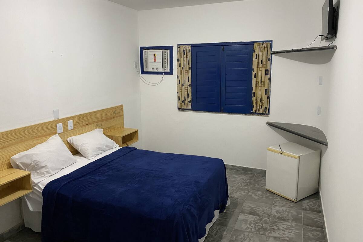 Ein kleines, sauberes Zimmer mit einem Einzelbett, einem Nachttisch, einer Kommode, einem Fenster mit Vorhängen und einem an der Wand befestigten Fernseher.