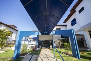 Um espaço ao ar livre coberto, com um caminho que leva à entrada de um edifício, cercado por vegetação e plantas em vasos.