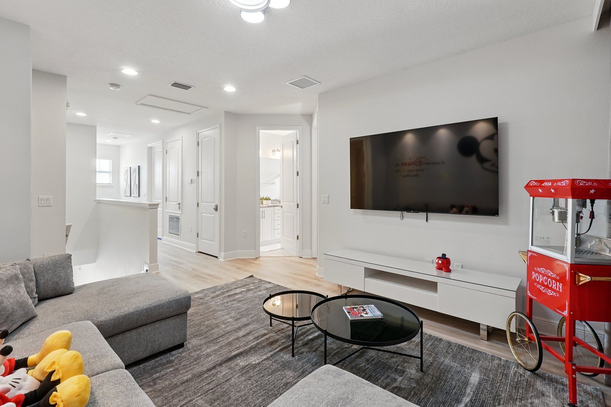 A modern living room with a grey sectional sofa, a glass coffee table, a flat-screen TV mounted on the wall, and a red popcorn cart.