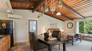A room with a wooden ceiling, a dining table with chairs, a bed, a window with a view of greenery, and a kitchen area with a refrigerator. - Cozy Up at a Nature Inspired Boutique Studio in the Trees - River Nearby (Bend)