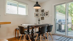 En matplats med ett träbord, svarta stolar och en svart hänglampa. På väggen hänger inramade tavlor, och en skjutdörr i glas leder ut.
