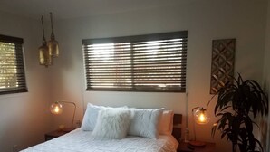 Ein ordentlich bezogenes Bett mit weißer, gesteppter Decke, zwei Nachttischlampen, einer Topfpflanze und einem Fenster mit Jalousien.