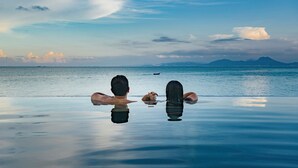 Un couple se détend au bord d’une piscine à débordement, avec vue sur la mer et les montagnes au loin.