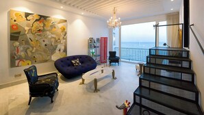 Un salon moderne avec un grand tableau abstrait, un fauteuil en forme de poire bleu, une chaise noire, un escalier et de grandes fenêtres donnant sur la mer.
