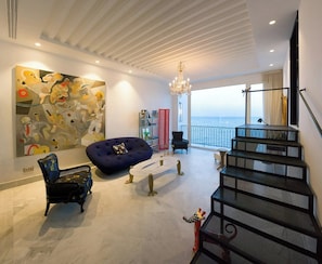 A modern living room with a large abstract painting, a blue bean bag, a black chair, a staircase, and a large windows overlooking the sea.