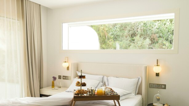 Une chambre d’hôtel avec un lit, des tables de chevet, un plateau avec des articles pour le petit-déjeuner, une fenêtre donnant sur un paysage verdoyant et des lampes fixées au mur.