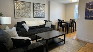 Een woonkamer met een donkergrijze bank, een salontafel, en een eethoek met een tafel en stoelen. Boven de bank hangen twee decoratieve wanddecoraties.