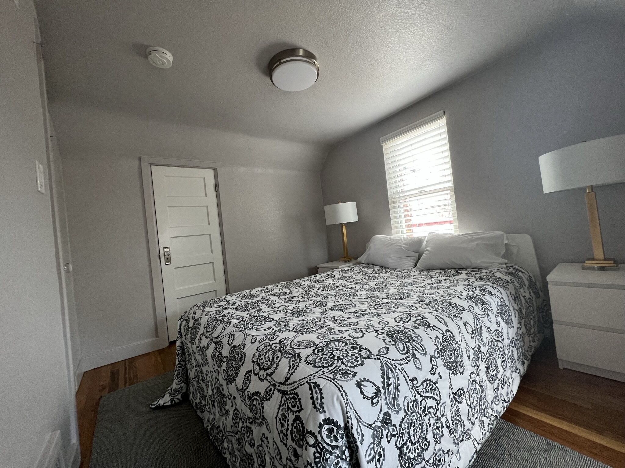 A bedroom with a bed, a nightstand, a lamp, a window with blinds, a door, and a ceiling light.