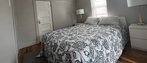 A bedroom with a bed, a nightstand, a lamp, a window with blinds, a door, and a ceiling light.