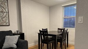 Een eethoek met een donker houten tafel en stoelen, een grijze bank met een gekleurd kussen, en een raam met zonwering.