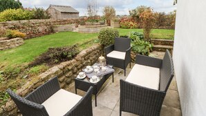 Un patio exterior con mobiliario de mimbre y una mesa lista para comer, rodeado de un jardín y un muro de piedra.