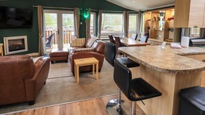 A modern living room with a sofa, a coffee table, and a dining area with chairs. There is a kitchen with a counter and cabinets. - Elder Lodge (Windermere)