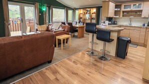 A modern interior with a brown leather sofa, a wooden dining table, and a kitchen area with wooden cabinets and a bar stool. - Elder Lodge (Windermere)