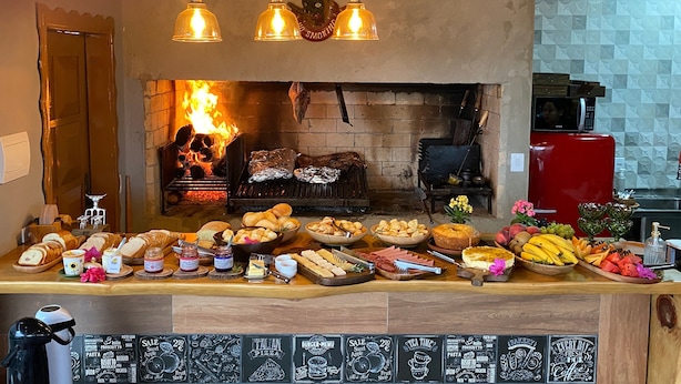 A restaurant with a brick fireplace, a variety of food displayed on the counter, and a coffee machine. - Refúgio dos Coiotes (Cambara do Sul)