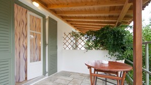 Un patio cubierto con techo de madera, suelo de piedra, una mesa con sillas y una puerta con contraventanas verdes.