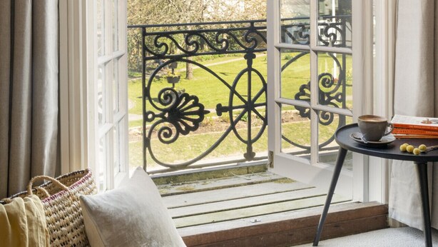 Una habitación con un ventanal amplio, un sofá, una mesita con una taza y libros, y vistas a un jardín.