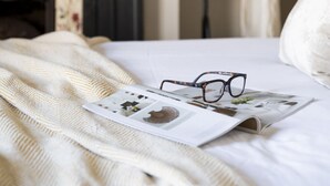 Una cama con una manta blanca, una revista y unos lentes encima.