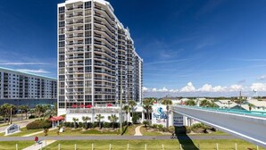 A multi-story hotel with a modern design, featuring balconies on each floor, a prominent sign for the Surfside Resort, and a view of the surrounding area including a road and greenery. - Beachfront Bar! Restaurant on Site! Spa! Tennis! Beach Service Included! (Miramar Beach)