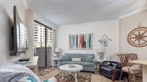 A modern living room with a blue sofa, a coffee table, a TV, and a dining area with metal chairs. - Beachfront Bar! Restaurant on Site! Spa! Tennis! Beach Service Included! (Miramar Beach)