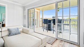A modern living room with a beige sofa, a patterned pillow, and a large sliding glass door leading to an outdoor patio with a dining table and chairs. - Condo with Golf Course Views | Near Attractions (Kissimmee)