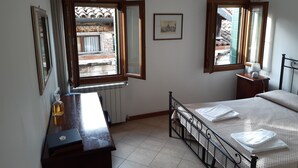 A bedroom with a bed, a desk, a radiator, and two windows with open curtains.