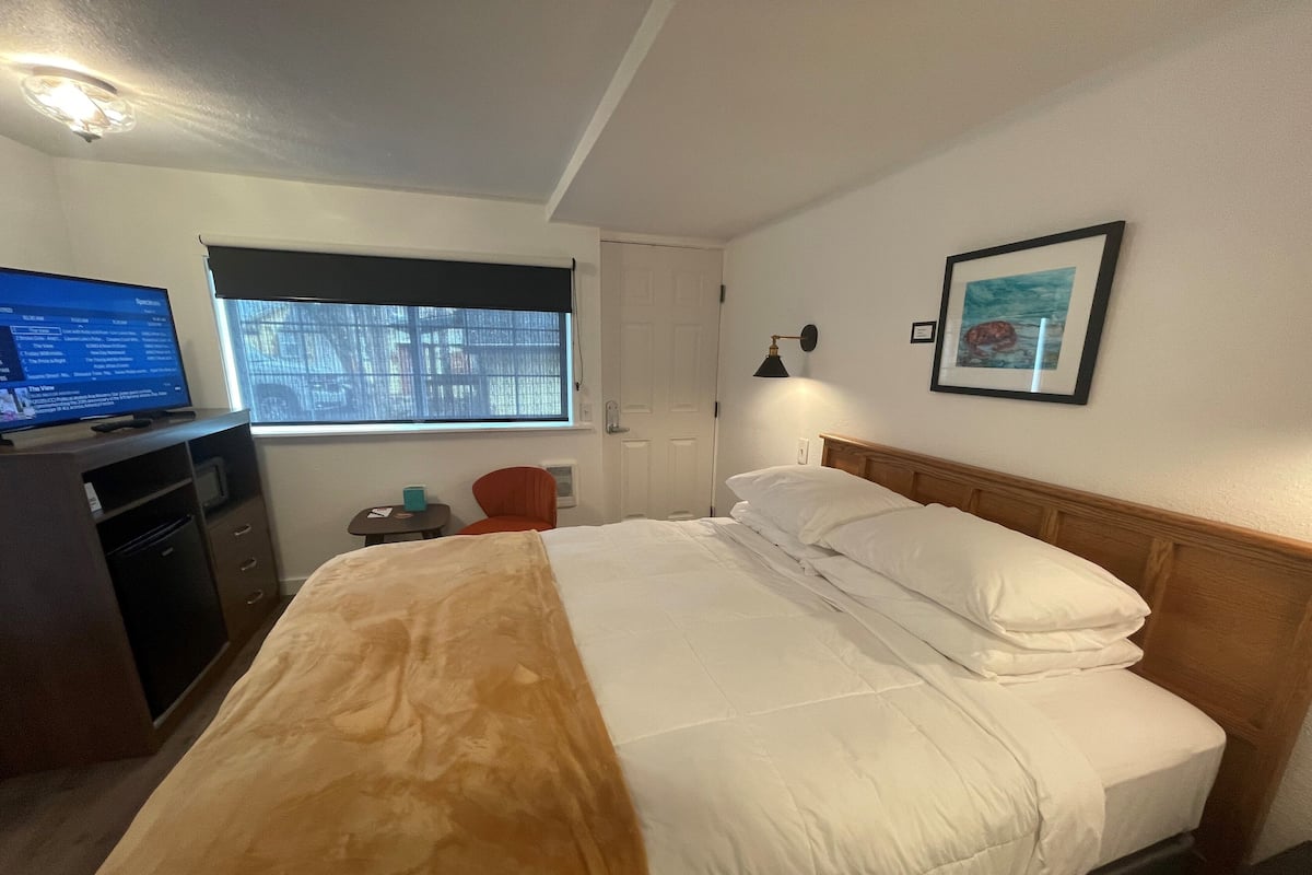 A hotel room with a bed, a desk with a computer, a chair, a window, and a framed picture on the wall.