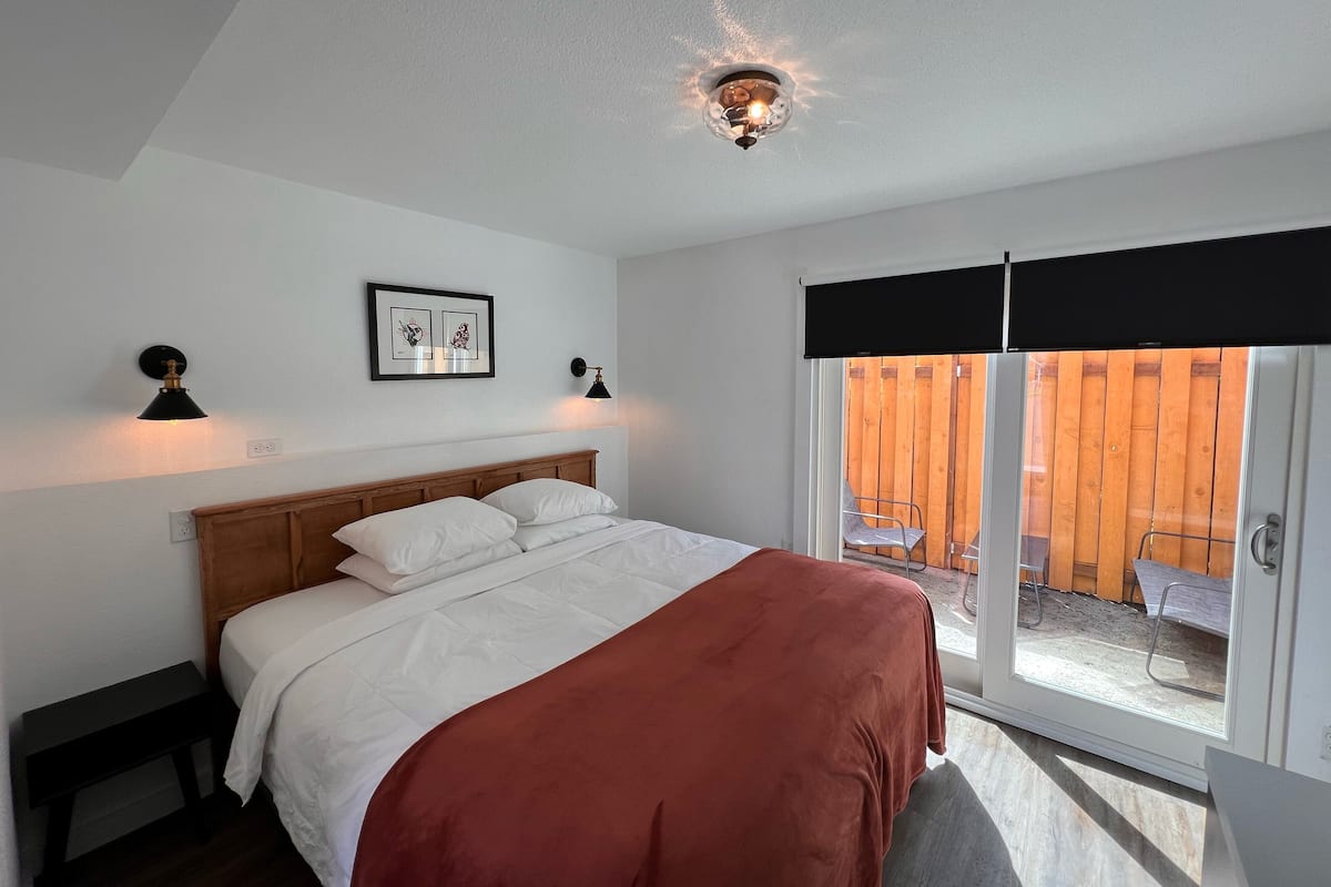 A bedroom with a bed, two bedside lamps, a ceiling light, a window with a view of a wooden fence, and a framed picture on the wall.