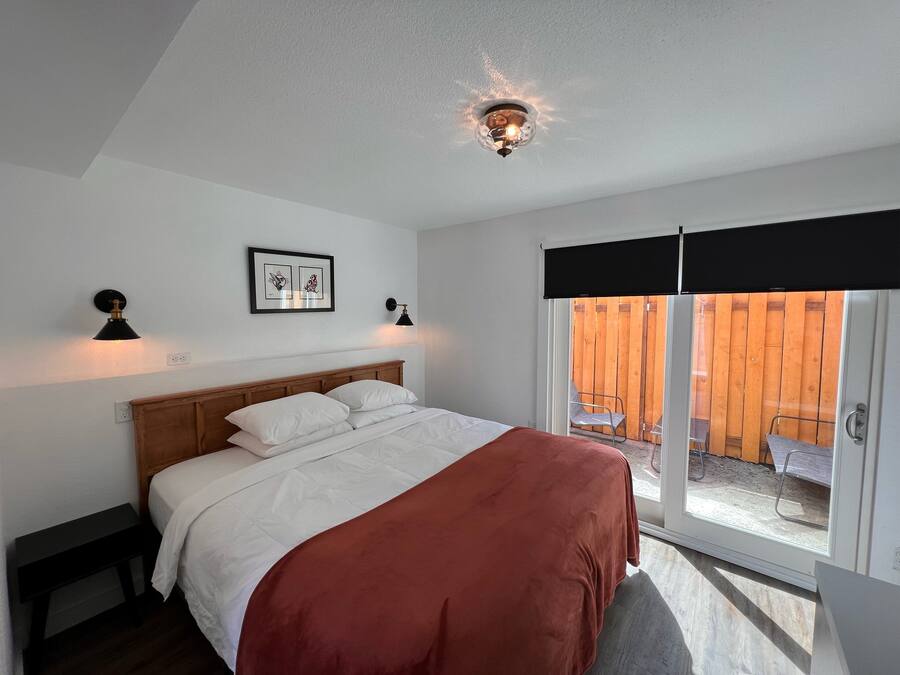 Ein Schlafzimmer mit einem Bett, zwei Nachttischlampen, einer Deckenleuchte, einem Fenster mit Blick auf einen Holzzaun und einem gerahmten Bild an der Wand.