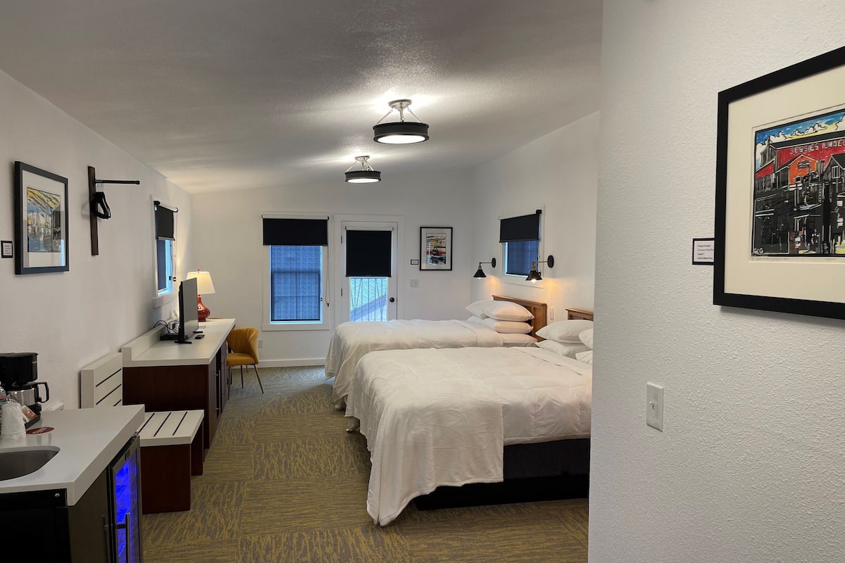 A hotel room with two beds, a desk, a chair, a small kitchenette, and a bathroom.