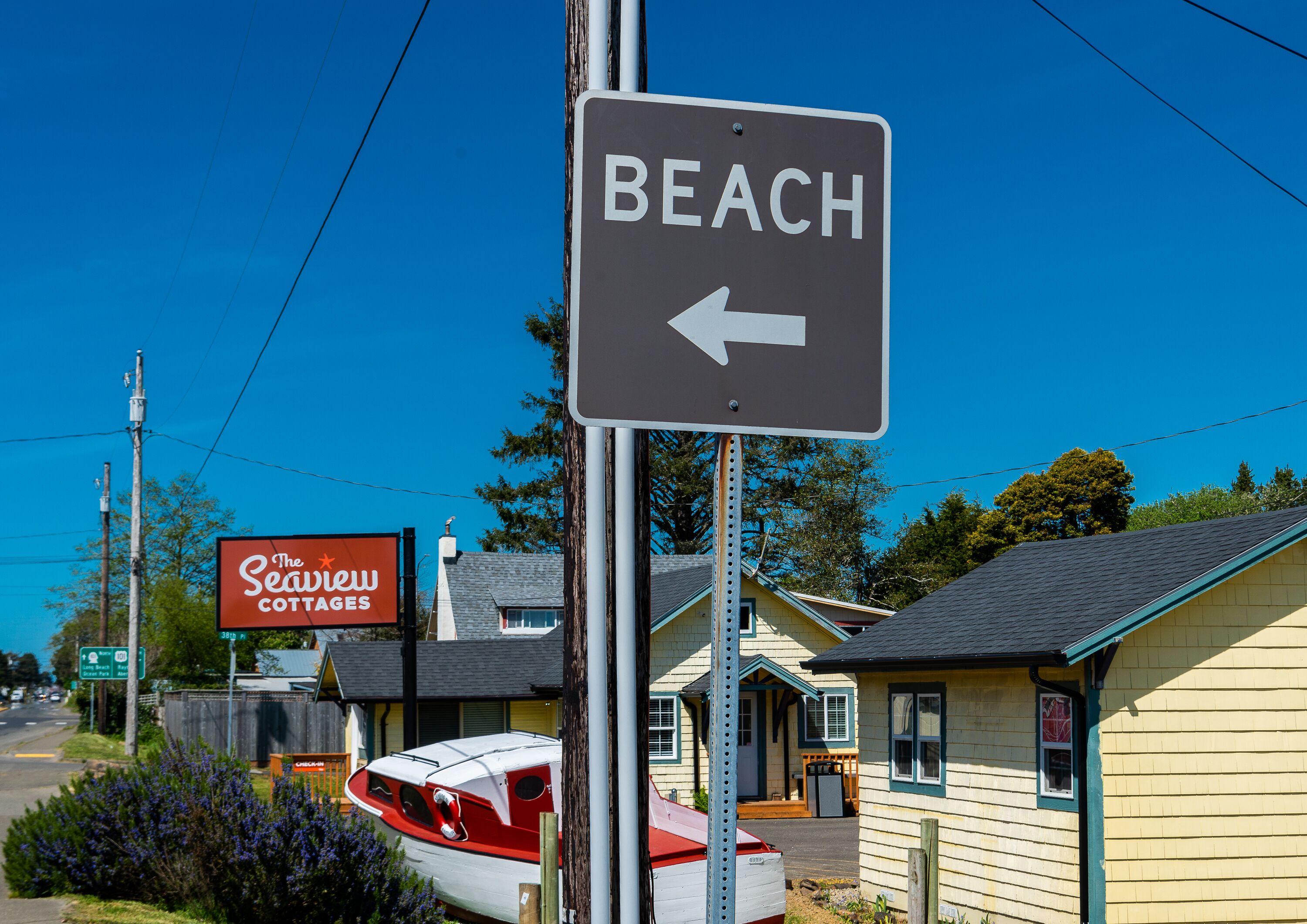 Un cartello da spiaggia con una freccia che indica a sinistra, un segnale rosso per The Seawell Cottages, e una barca parcheggiata di fronte a un edificio giallo.