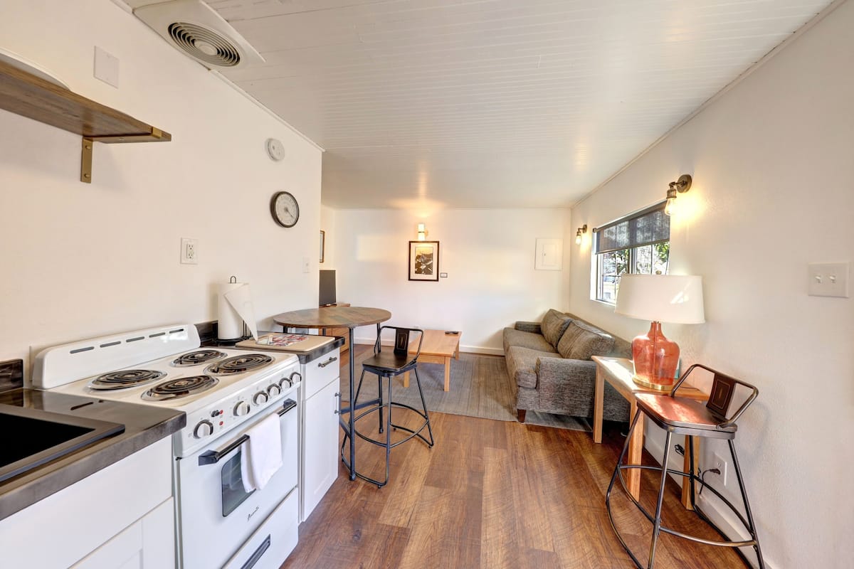 A compact kitchen with a stove, oven, and bar stools. There is a dining table and a sofa in the living area.