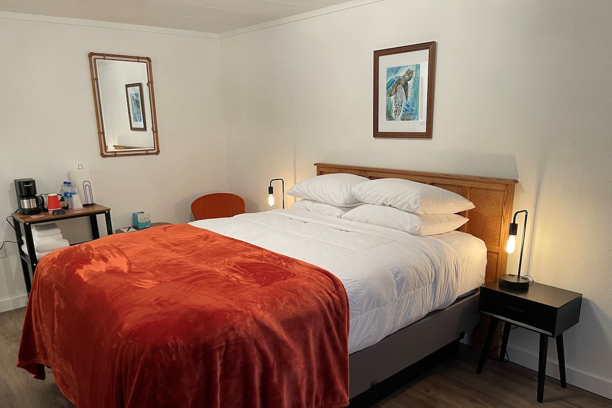 A hotel room with a bed, a nightstand, a chair, a desk with a coffee maker, and framed artwork on the walls.