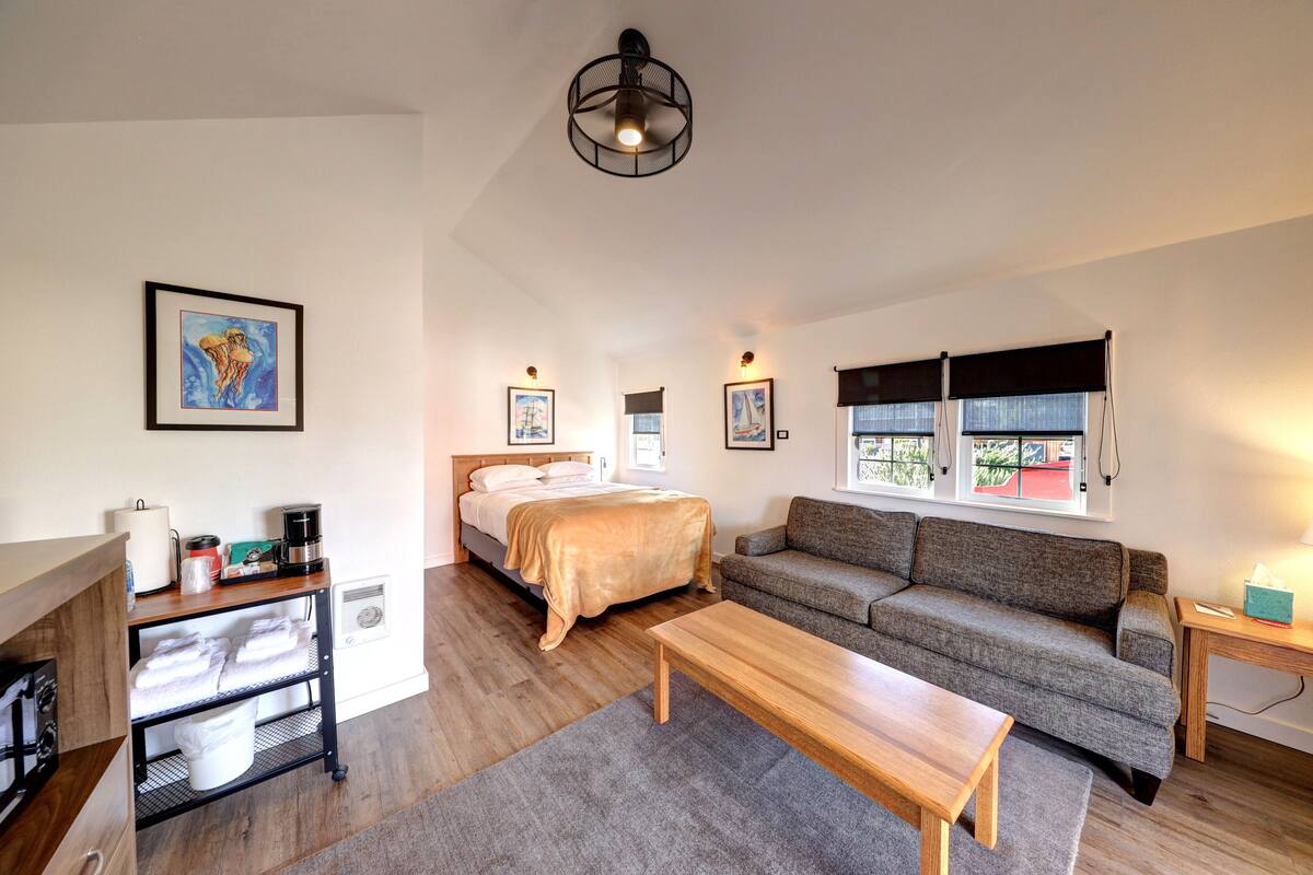 A modern, well-lit room with a bed, sofa, and a wooden table. There are framed pictures on the walls, a window with curtains, and a ceiling light.