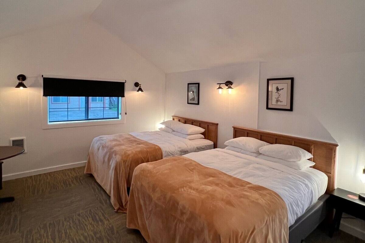 A room with two beds, a ceiling fan, a window with blinds, wall-mounted lights, and a small table.