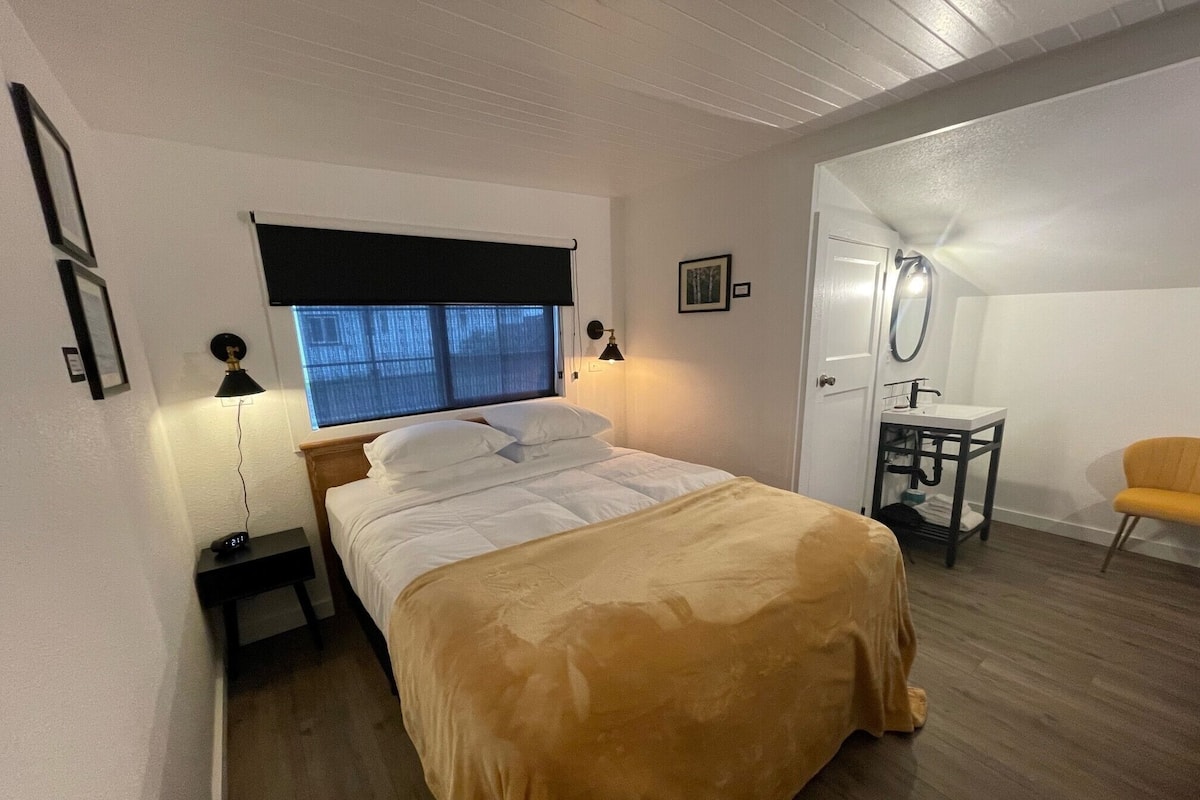 A bedroom with a bed, bedside lamps, a window with blinds, a small table with a sink, and a chair.