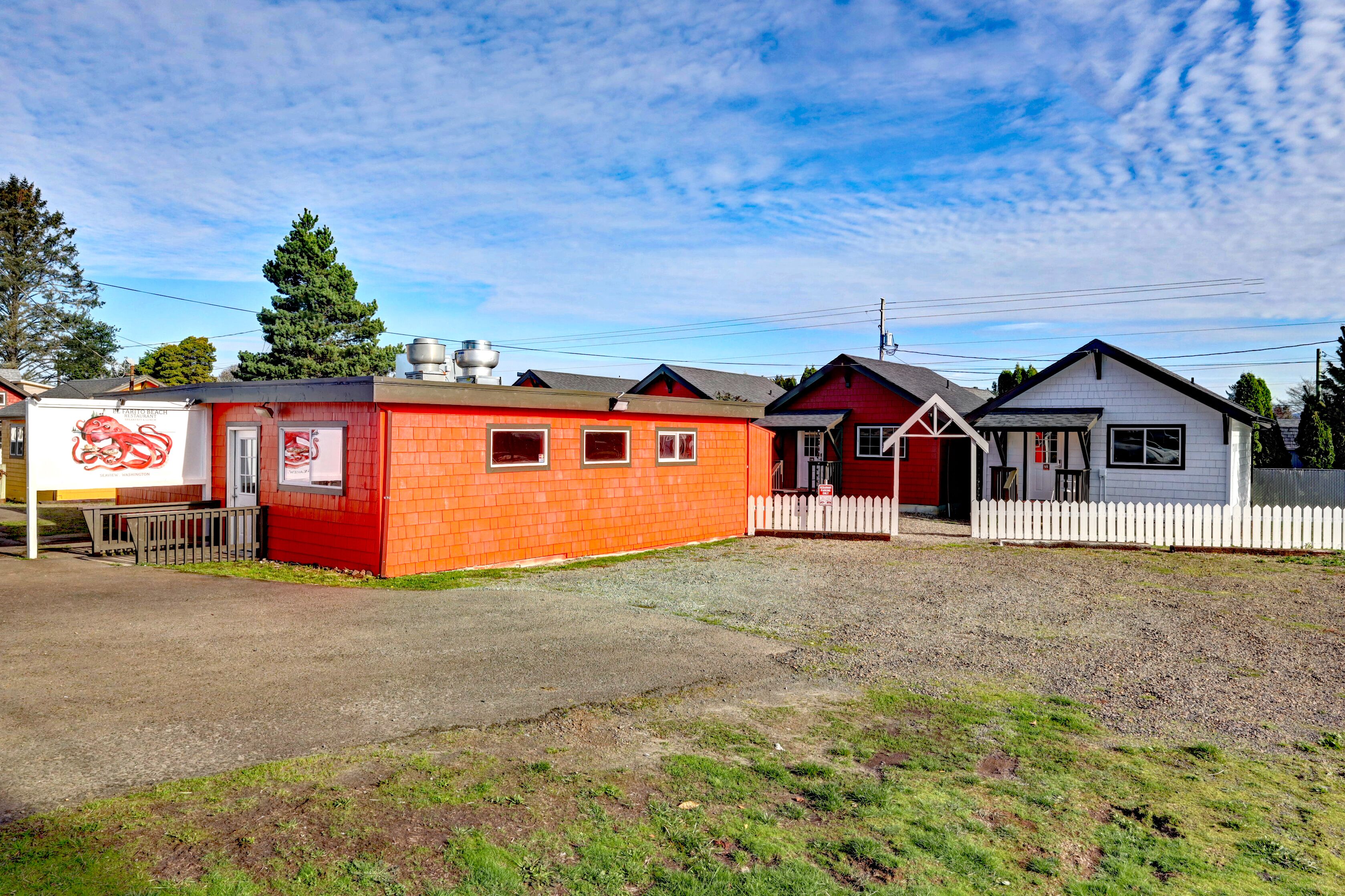 A row of small, single-story buildings with a gravel driveway and a white picket fence.
