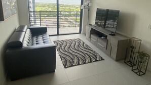 A modern living room with a sofa, a patterned rug, a TV on a wooden cabinet, and a balcony with a view of the city. - Beautiful Condo at Downtown Doral! (Doral)