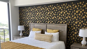 A large bed with a wooden frame, white bedding, and a patterned throw blanket. There are two bedside tables with lamps, and a wall with a circular pattern. - Beautiful Condo at Downtown Doral! (Doral)