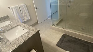 A modern bathroom with a granite countertop sink, a toilet, a glass-enclosed shower, and a glass door leading to a shower area. - Beautiful Condo at Downtown Doral! (Doral)