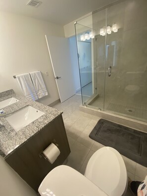 A modern bathroom with a granite countertop sink, a toilet, a glass-enclosed shower, and a glass door leading to a shower area. - Beautiful Condo at Downtown Doral! (Doral)