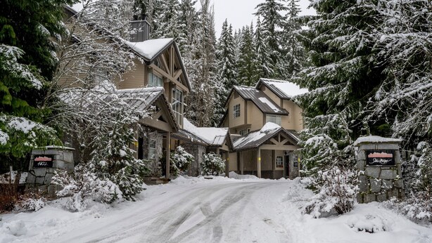 Ein mit Schnee bedecktes Chalet mit Steineingang, mehreren Fenstern und einer verschneiten Auffahrt.