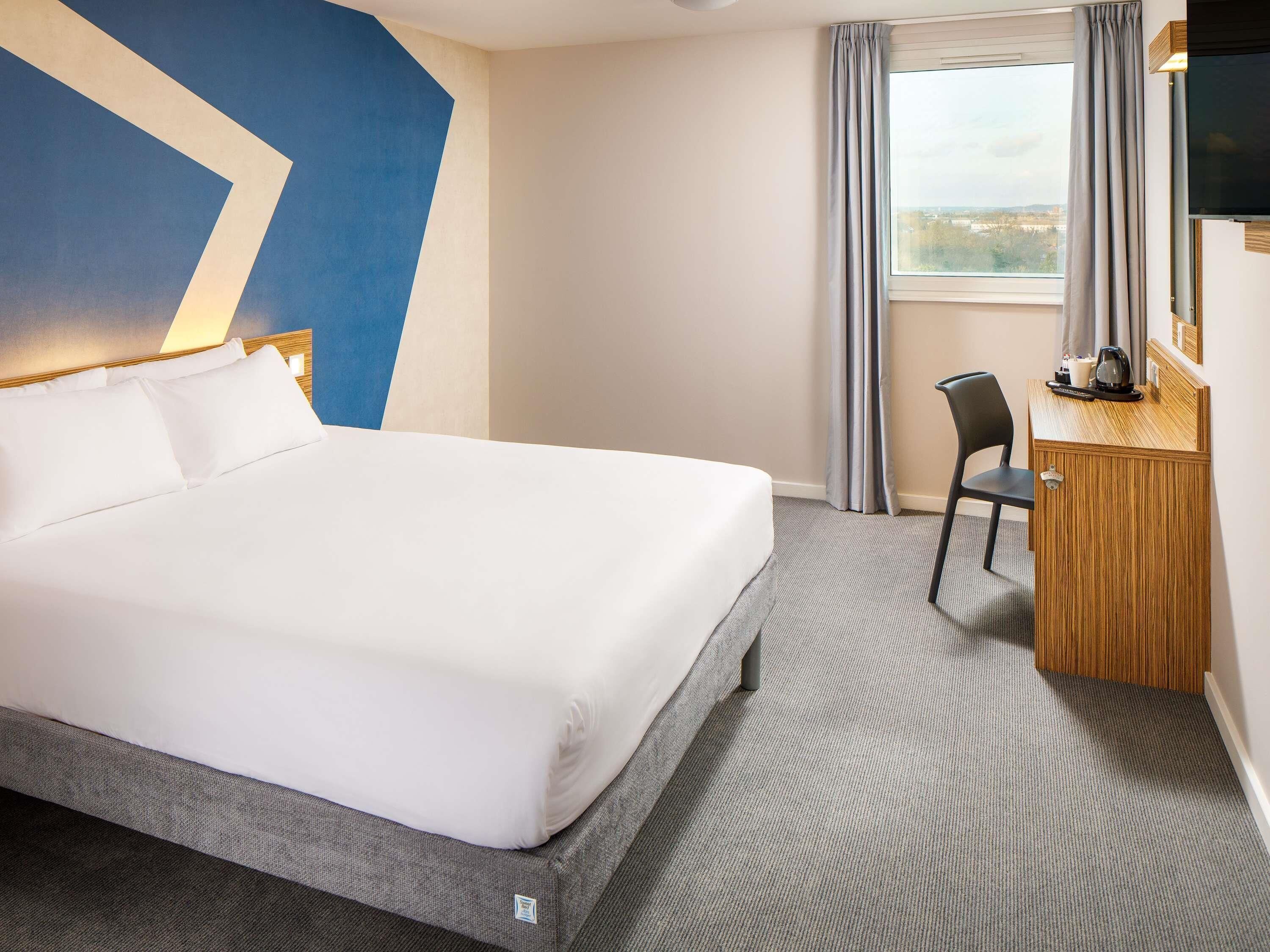 Una habitación de hotel con cama, escritorio, silla, televisión y ventana con cortinas.