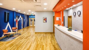 A reception area with a wooden floor, a counter with a clock, and a bar cafe sign in the background. - Ibis budget London Heathrow Central (Hounslow)