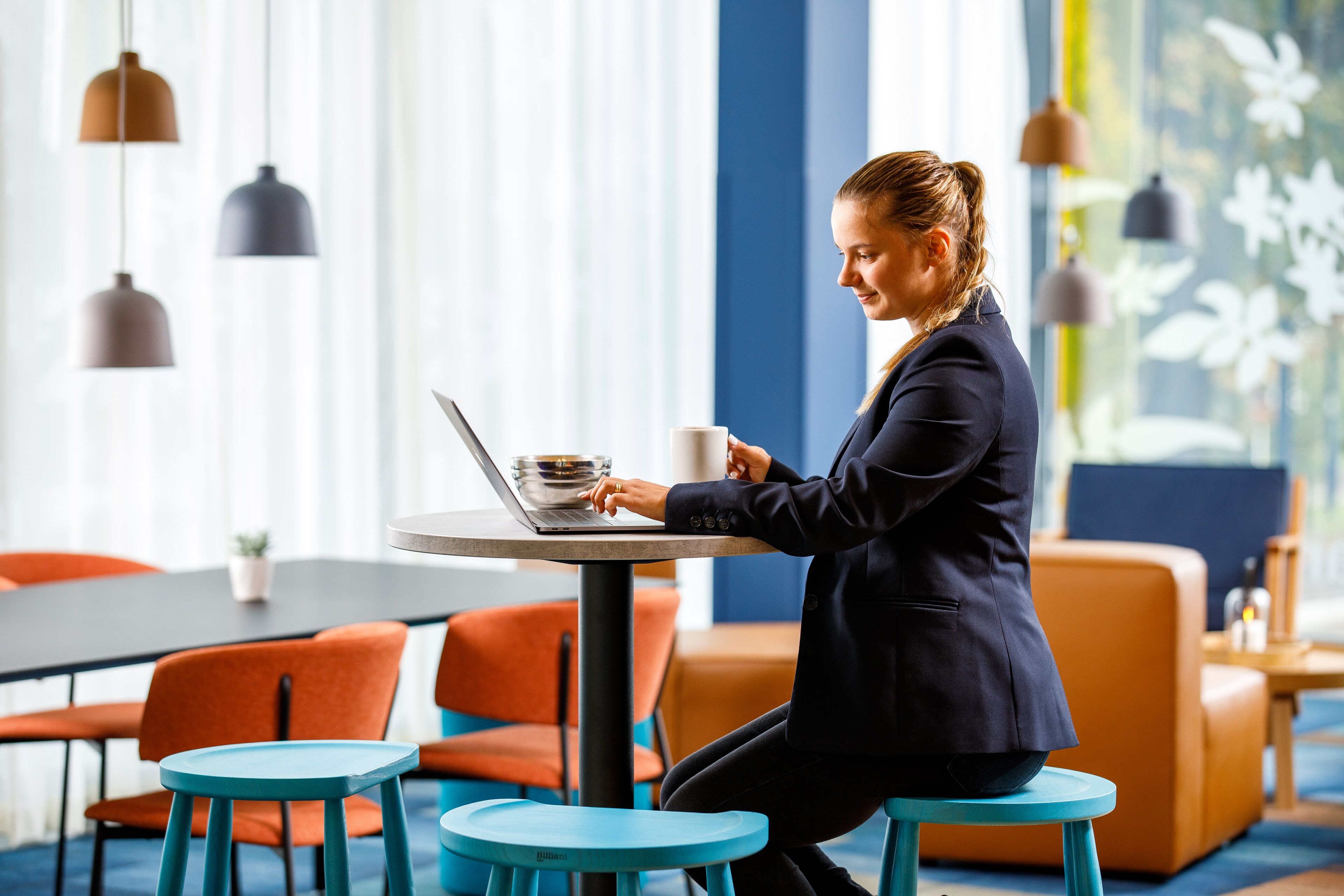 A person working on a laptop at a round table with a coffee cup, surrounded by orange chairs and blue stools in a bright room with large windows and pendant lights.