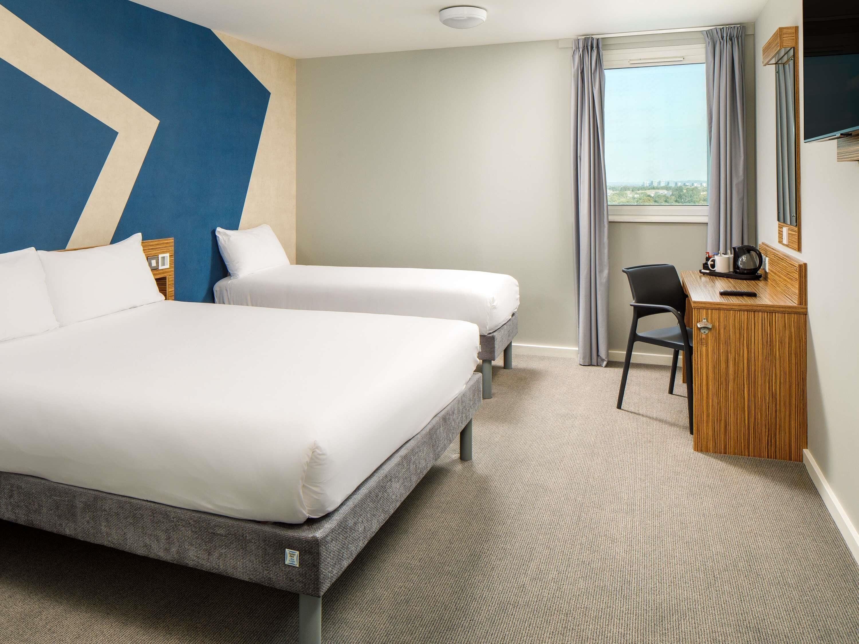 A hotel room with two beds, a desk, a chair, and a large window with curtains.