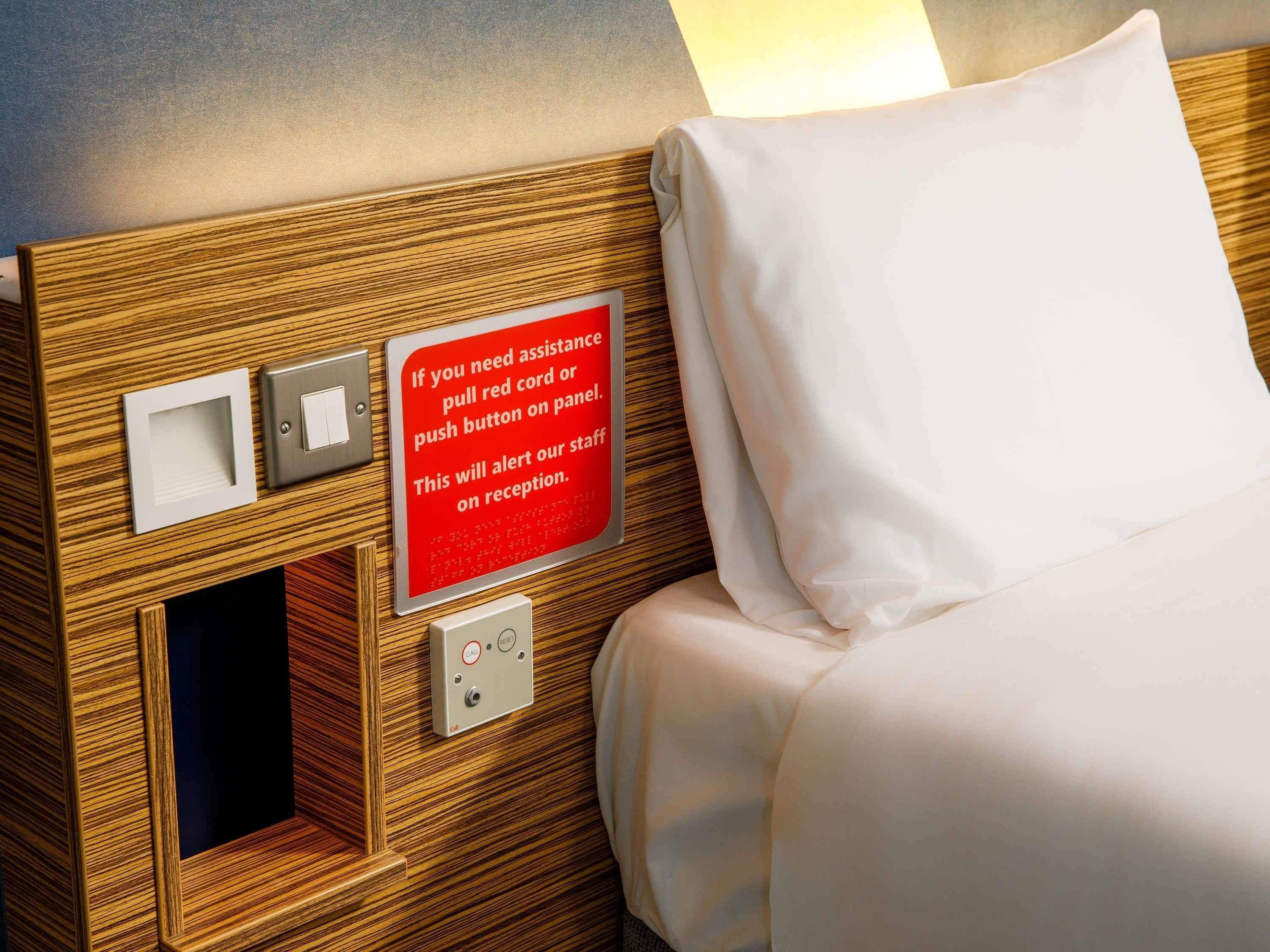 A hotel room with a wooden headboard, a bed with white bedding, a red assistance sign, a light switch, and a panel with a red cord.