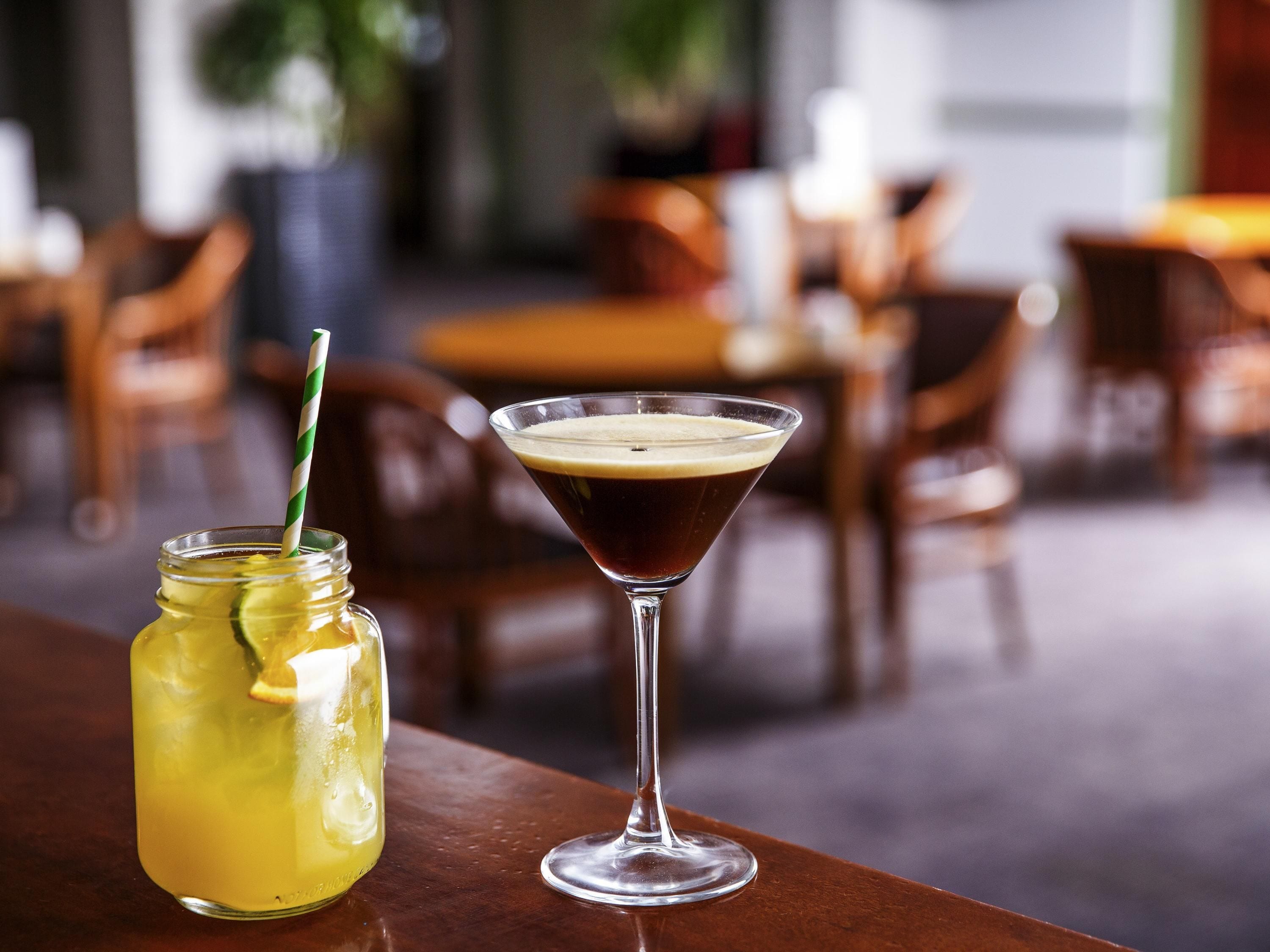 A martini glass and a mason jar drink on a wooden table in a restaurant setting.